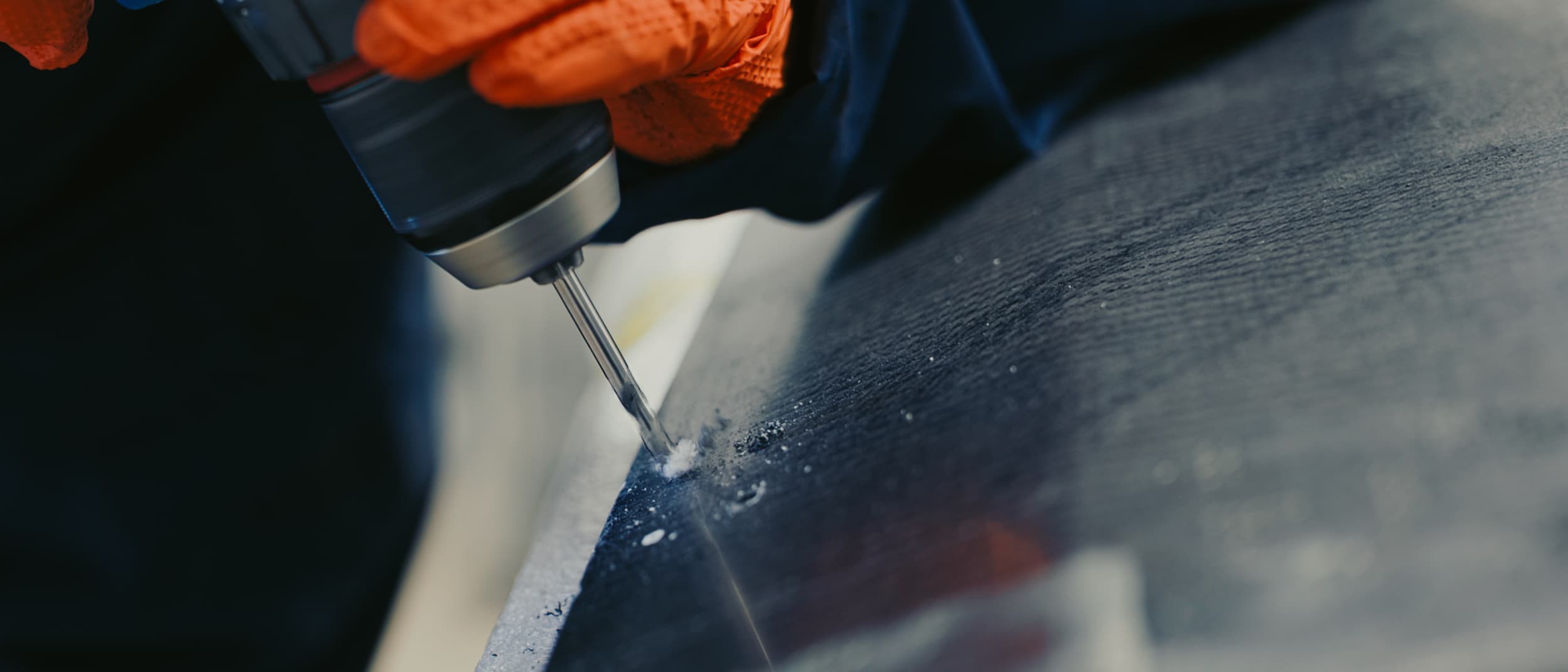 A gloved hand using a drill to finish the edge of a carbon fibre panel in the workshop.